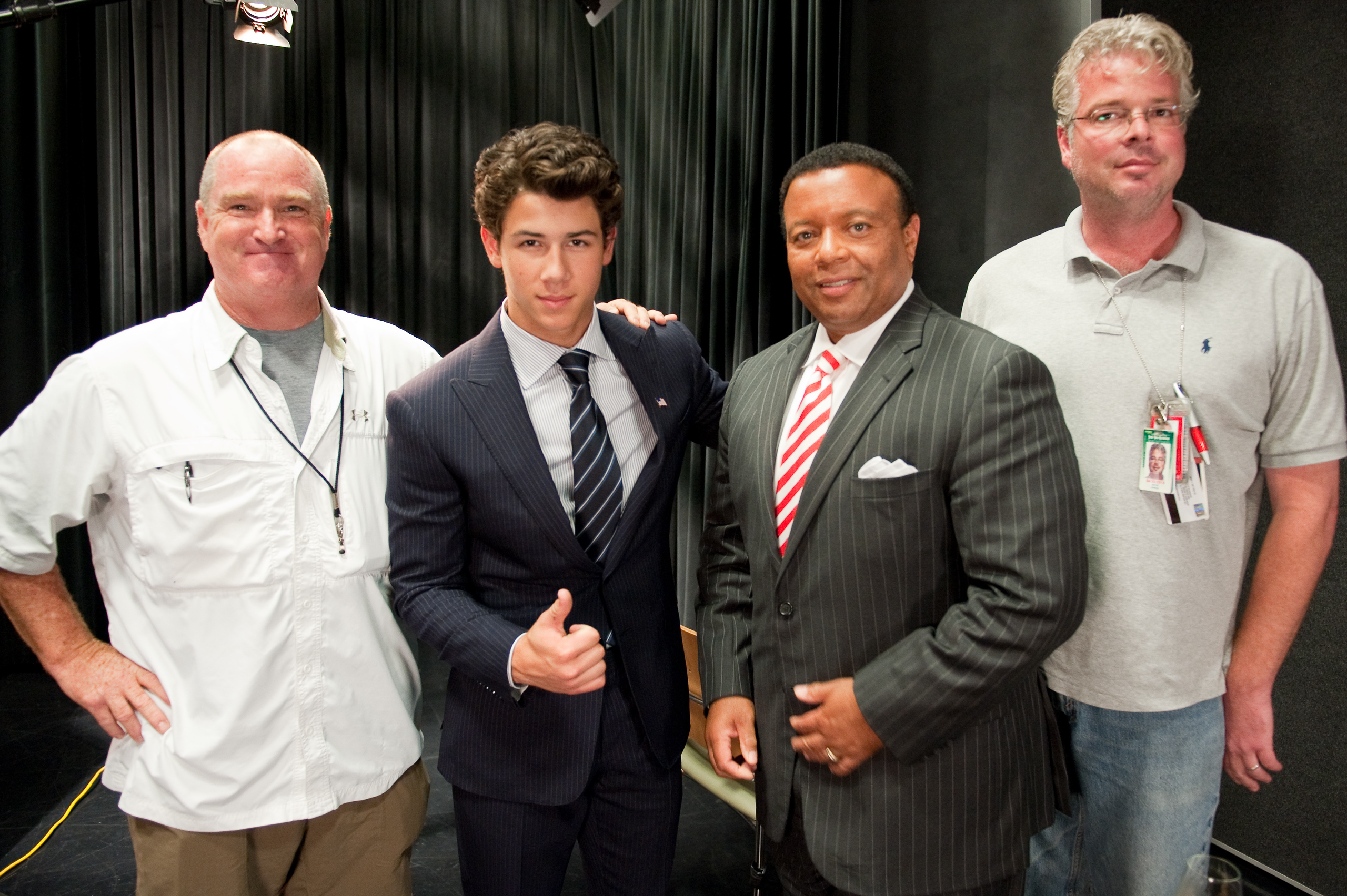 Four men pose for an epic portrait indoors; two wear business suits, one gives a thumbs up, and two sport casual clothing. Reminiscent of Nick Jonas’s style, they stand confidently against a black curtain backdrop for their group photo.