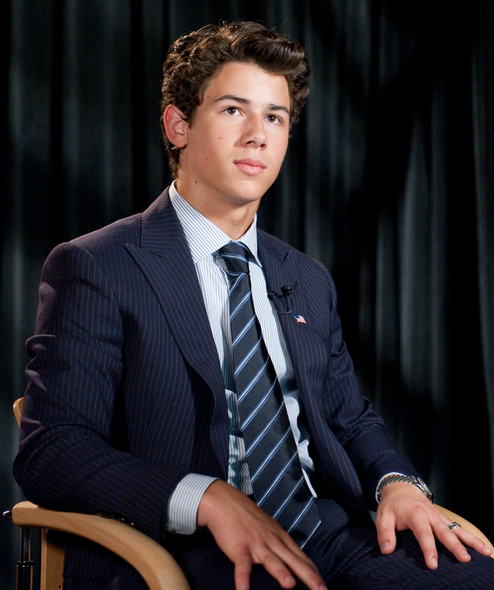 A young man in a pinstripe suit and striped tie sits on a chair with a backdrop of dark curtains, looking slightly to the side in this epic portrait reminiscent of Nick Jonas's classic style.