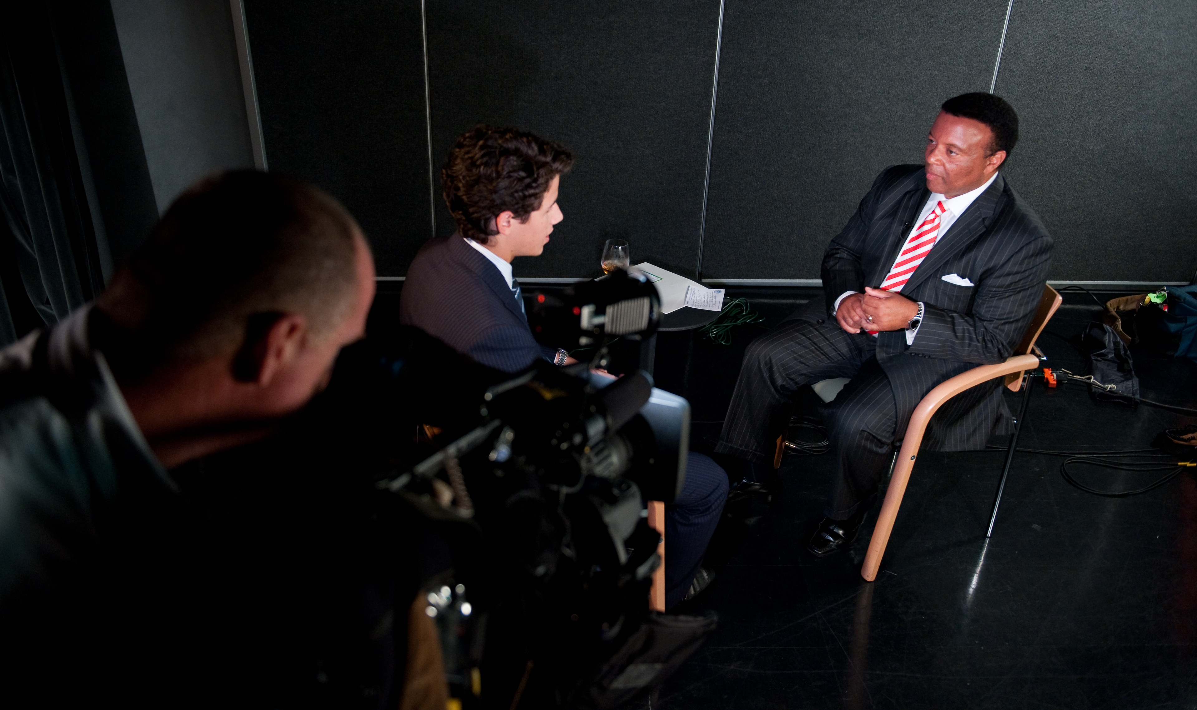 A man in a suit is being interviewed by another man while a cameraman films the conversation in a studio setting, creating an epic portrait reminiscent of celebrity interviews with stars like Nick Jonas.