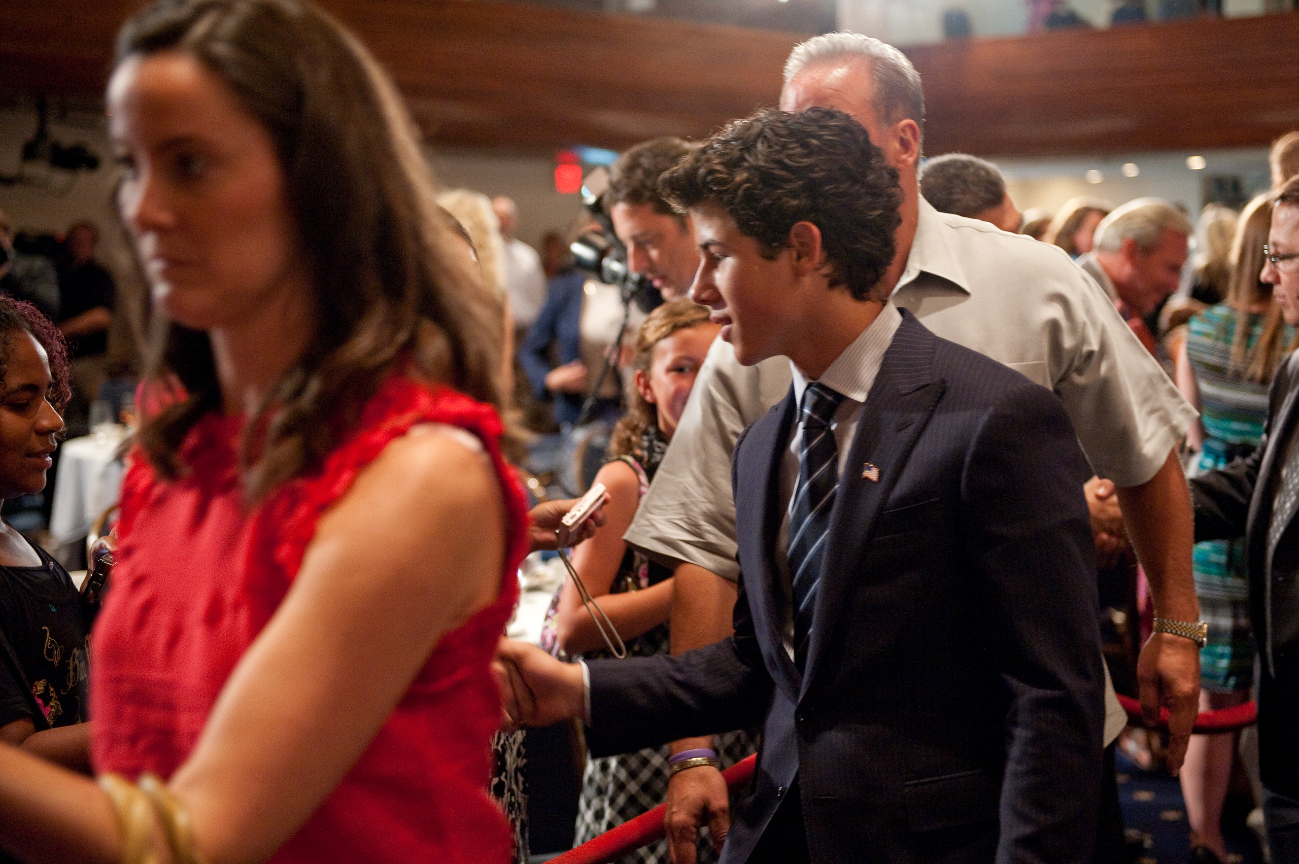 Nick Jonas, dressed in a sharp suit, walks past a crowd in a busy indoor setting while others stand nearby with microphones or cameras—an epic portrait of modern celebrity life.