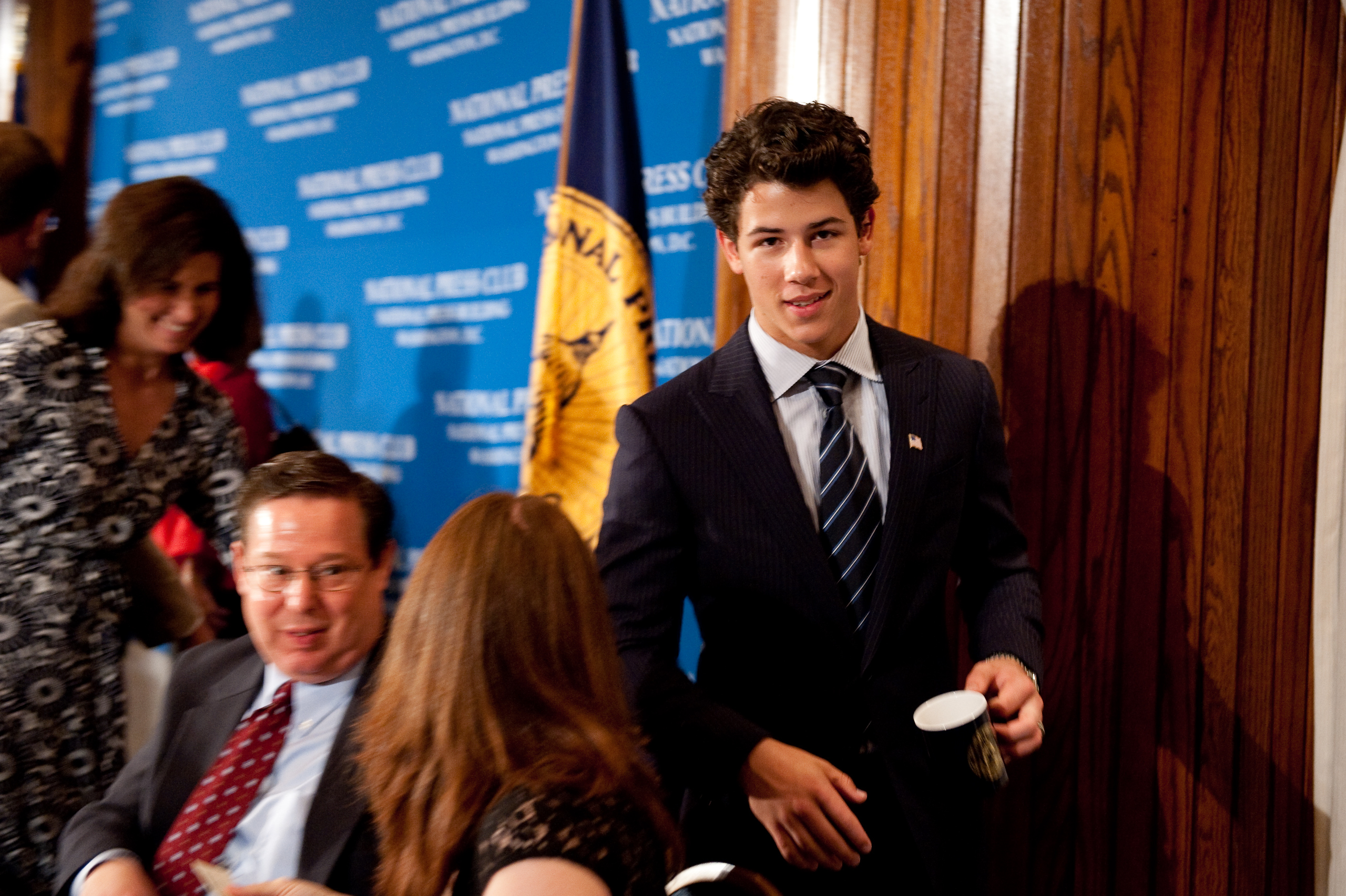 A young man in a suit, resembling Nick Jonas, holds a cup and stands near seated people at an event with an Epic Portrait vibe and a blue National Press Club backdrop.