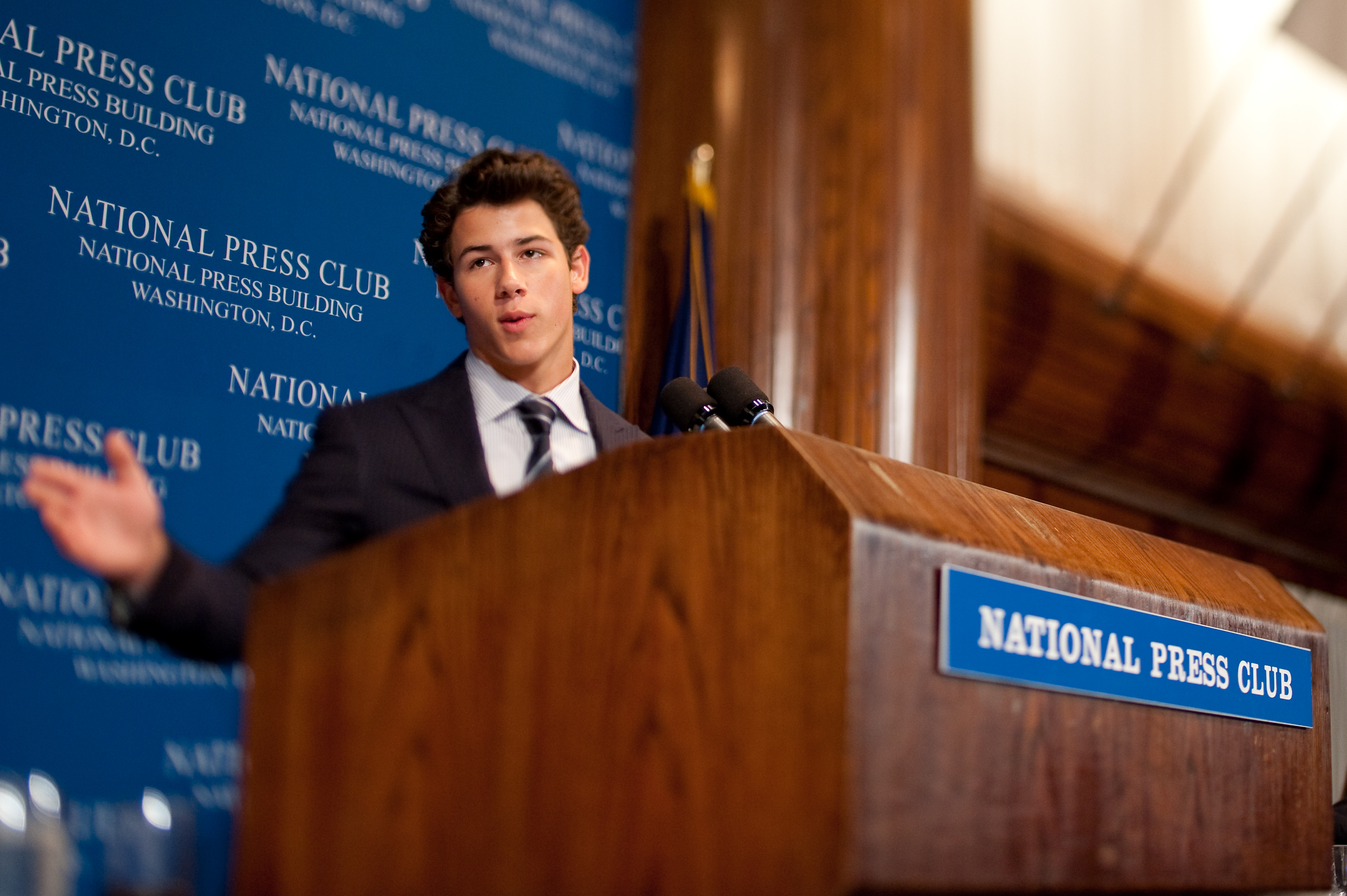 A man in a suit delivers an epic portrait of leadership as he speaks at a podium labeled "National Press Club," set against a blue backdrop in Washington, D.C.
