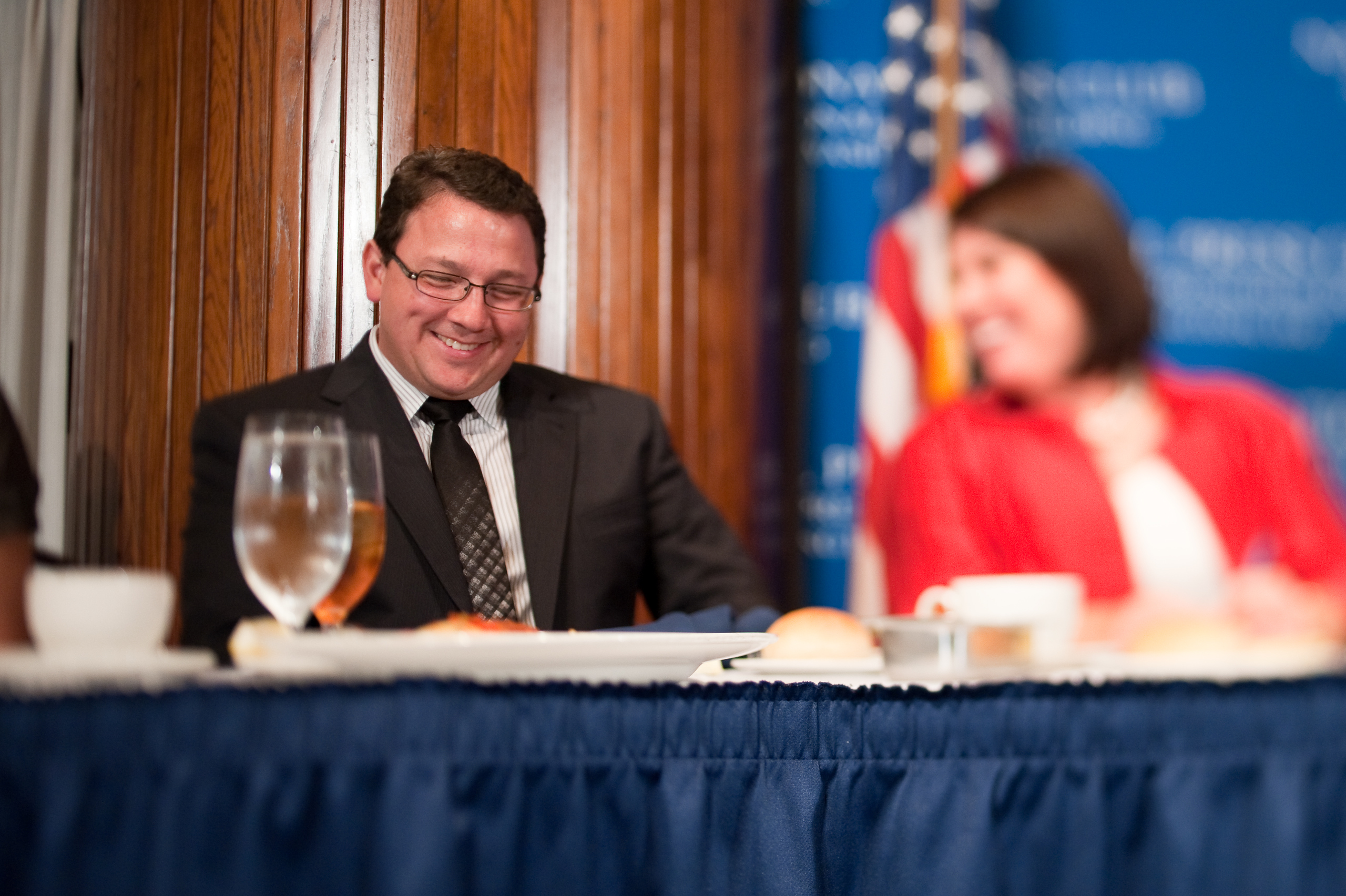 A man in a suit and tie sits at a table with food and water, smiling in an epic portrait next to a woman in red with a blurred background and a U.S. flag behind them. The scene evokes the style of Nick Jonas at an elegant event.