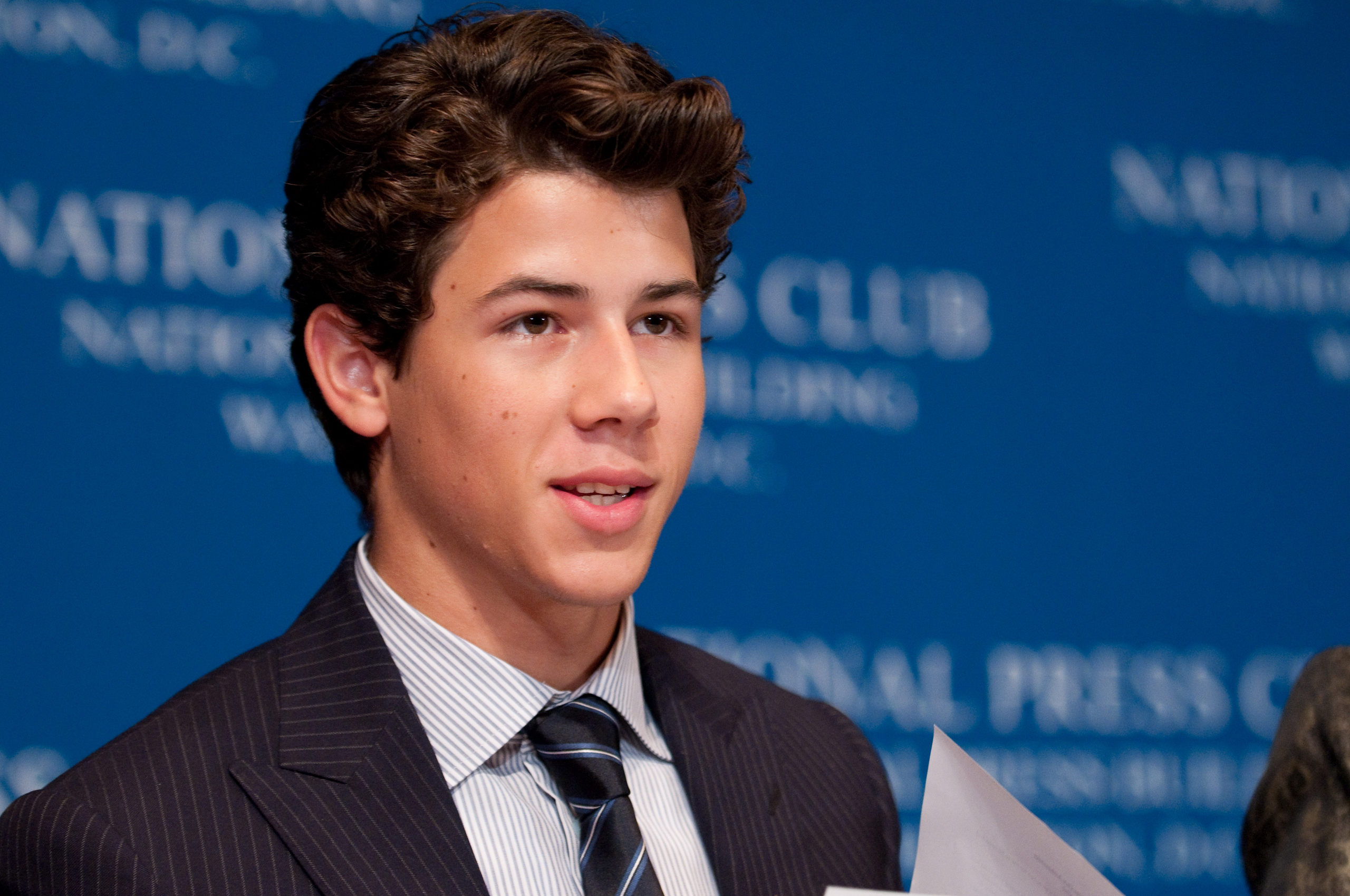 A young man in a suit and tie stands in front of a blue background with white text, holding a piece of paper and looking slightly to the side, reminiscent of an epic portrait of Nick Jonas.