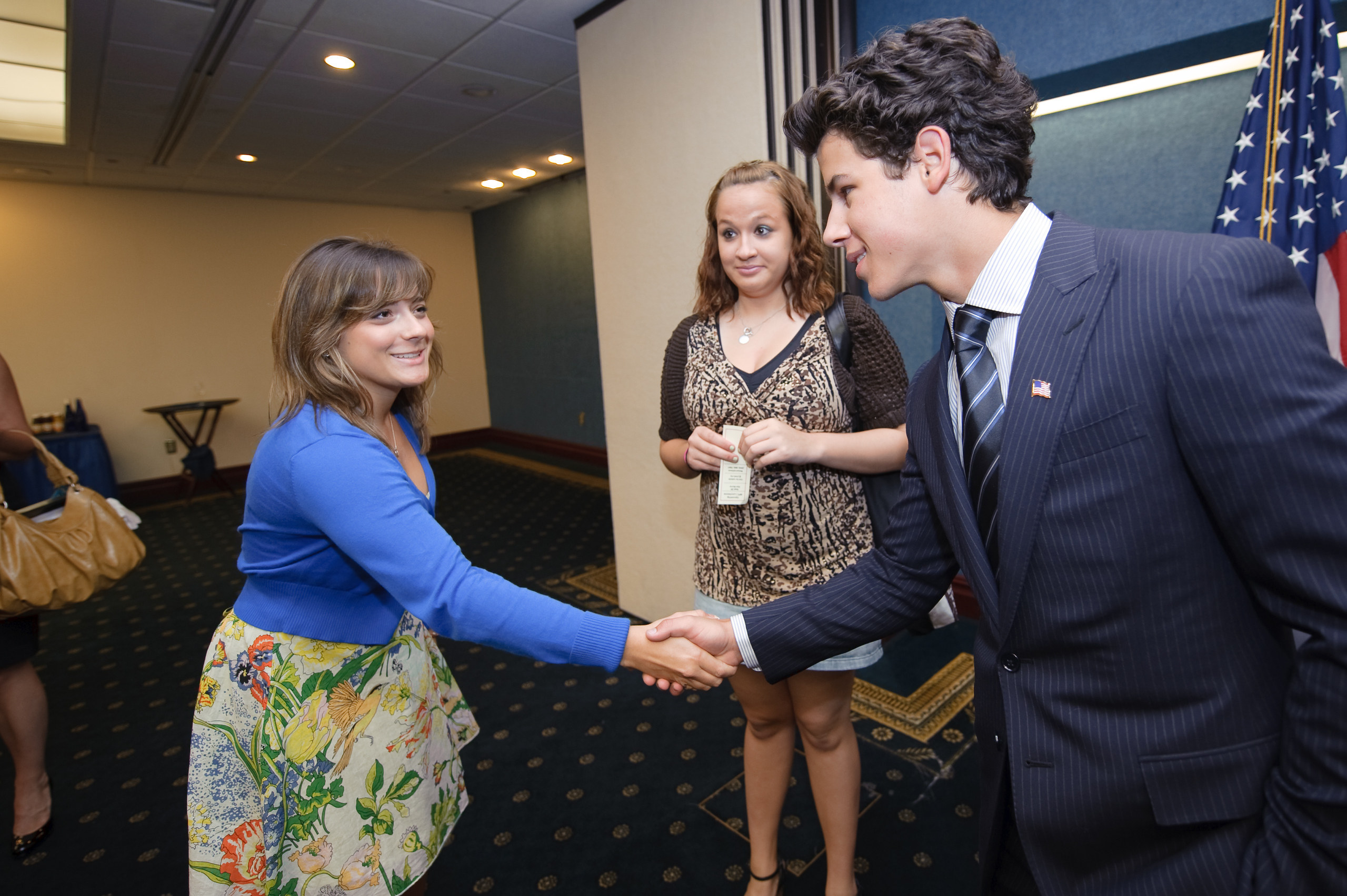 A woman in a blue top and floral skirt shakes hands with a man in a suit, while another woman stands nearby holding a card—an epic portrait reminiscent of Nick Jonas’s charismatic style.