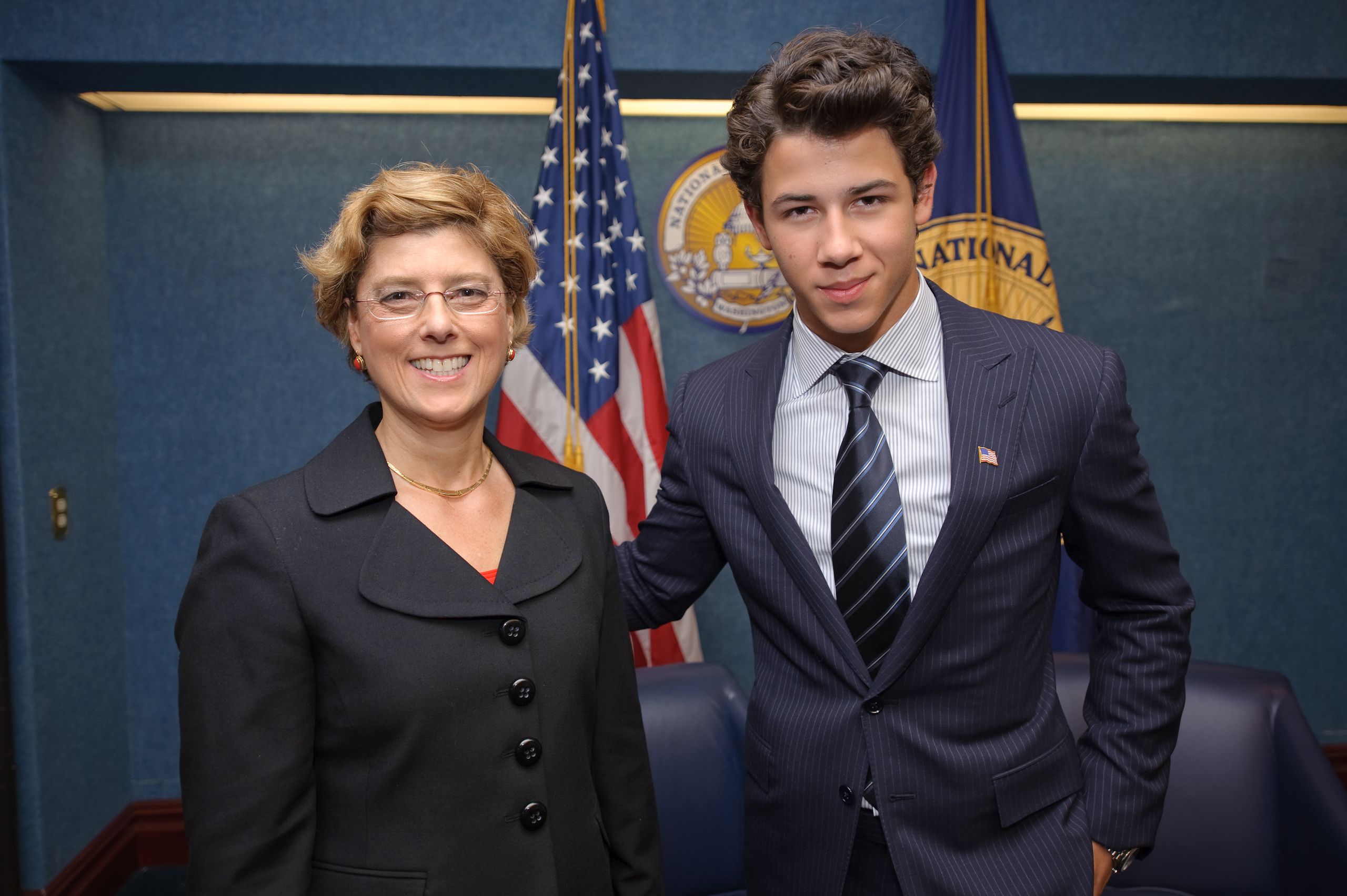 Two people in business attire pose together indoors in front of U.S. flags and official emblems, creating an epic portrait reminiscent of a Nick Jonas photoshoot.