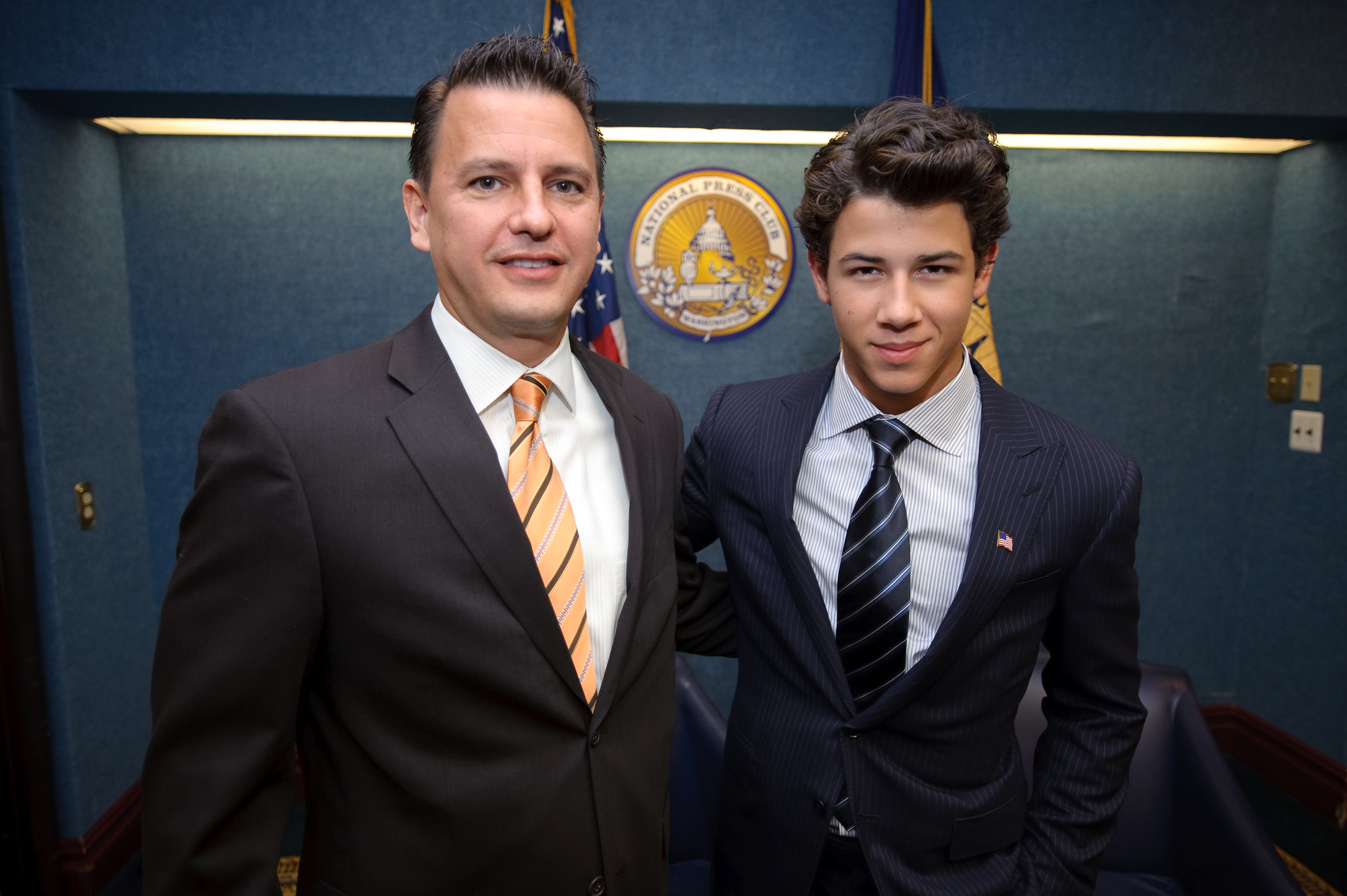 Two men in suits stand together in an office setting with a blue background and a government seal behind them, captured in an epic portrait style reminiscent of Nick Jonas’s confident presence.