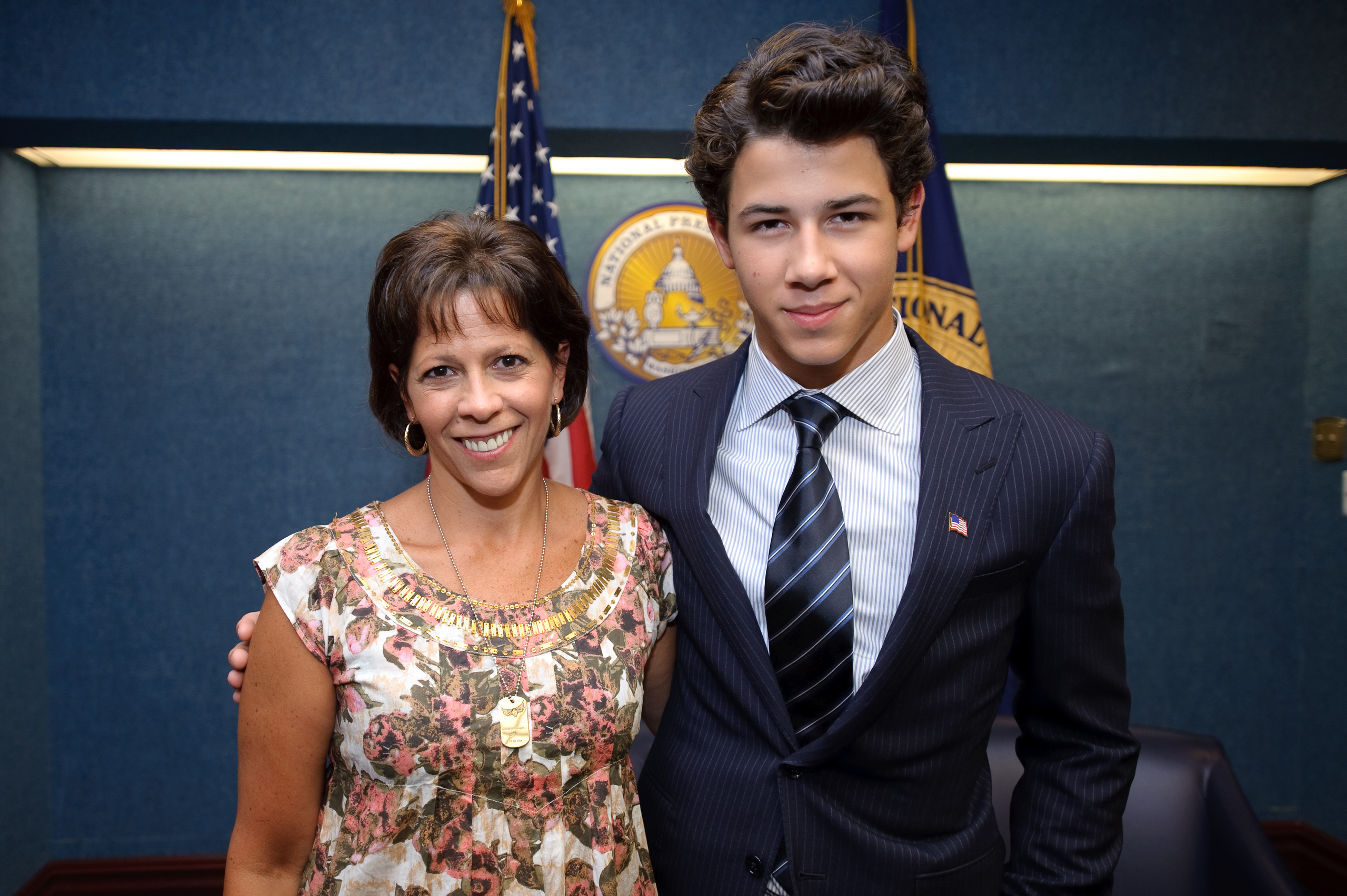 A woman in a floral blouse stands next to a man in a suit and tie in front of U.S. and state flags, creating an epic portrait against a blue wall with official seals.