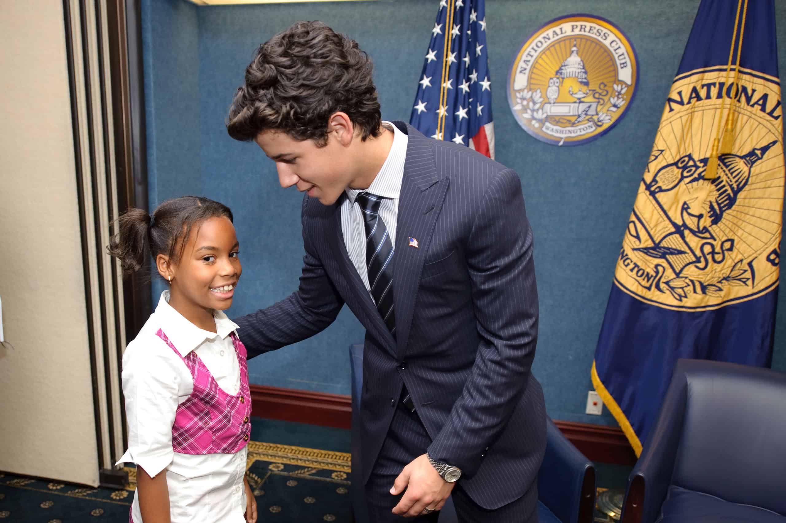 Epic Portrait: A man in a suit smiles and talks to a young girl in a pink plaid vest—reminiscent of Nick Jonas—at the National Press Club, with American and club flags waving proudly behind them.