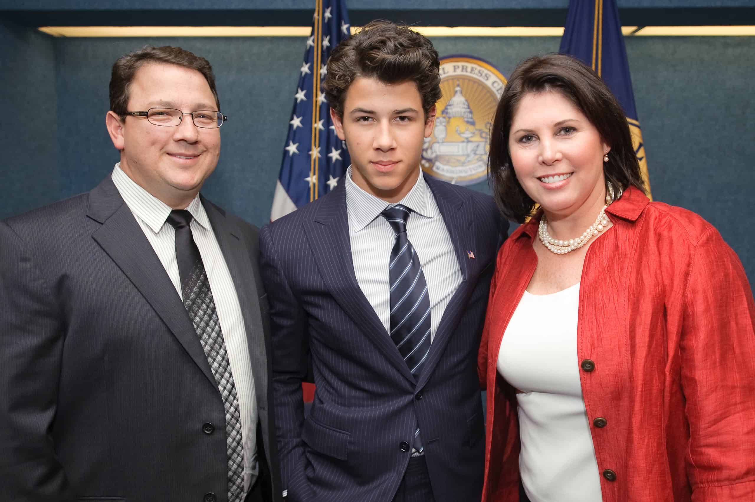 Three people in formal attire stand together indoors, creating an epic portrait in front of American flags and an official seal.