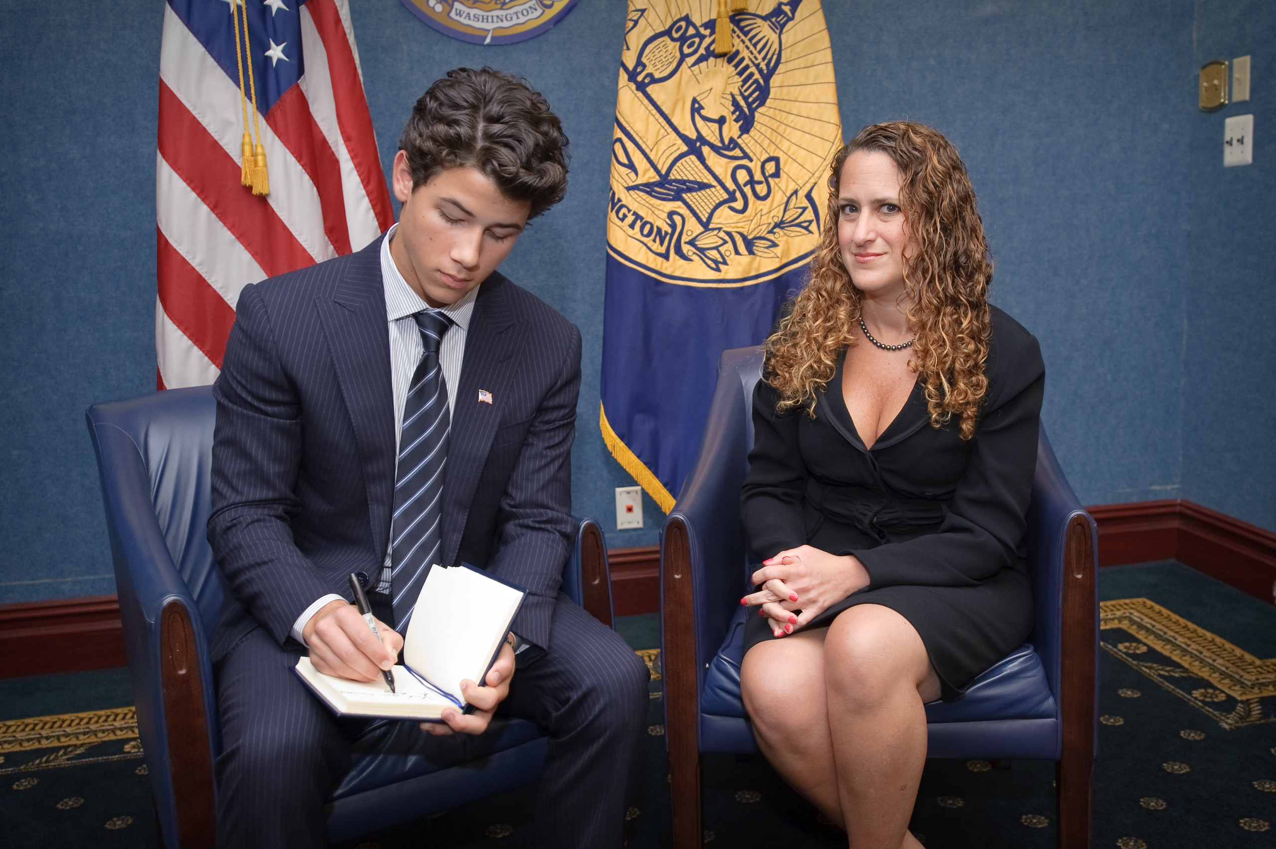 A man in a suit writes in a notebook beside a woman in a black dress; both pose for an epic portrait in an office setting adorned with flags, official emblems, and a touch of Nick Jonas style.