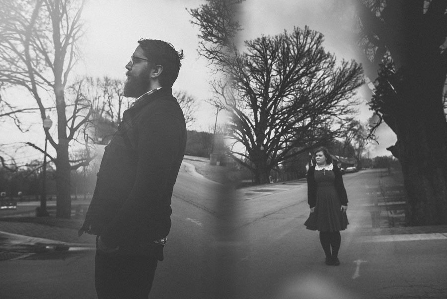Black and white photo of a bearded man in profile on the left and a woman standing on a road on the right, separated by a prisming blurred line.