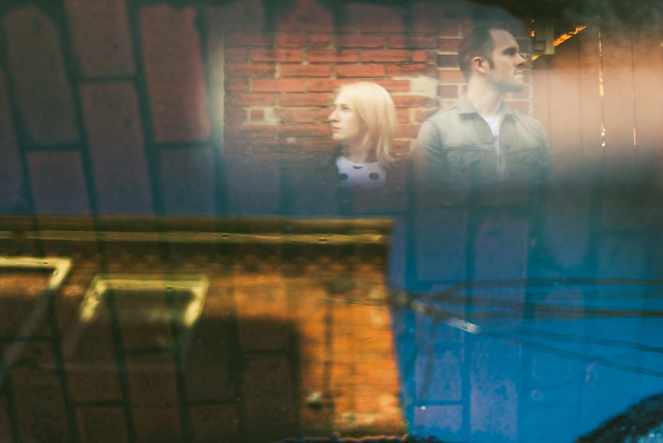 A man and a woman reflected in a puddle against a brick wall, with an overlay of golden light streaks created through the photography technique of prisming.