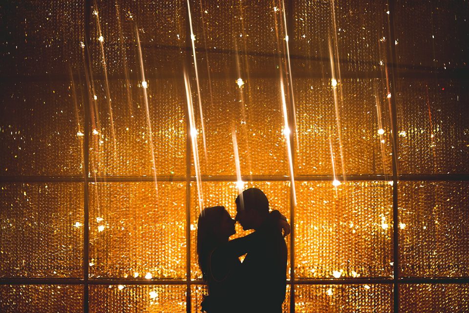 A silhouette of a couple kissing, backlit by a wall of golden twinkling lights, creating a prisming effect.