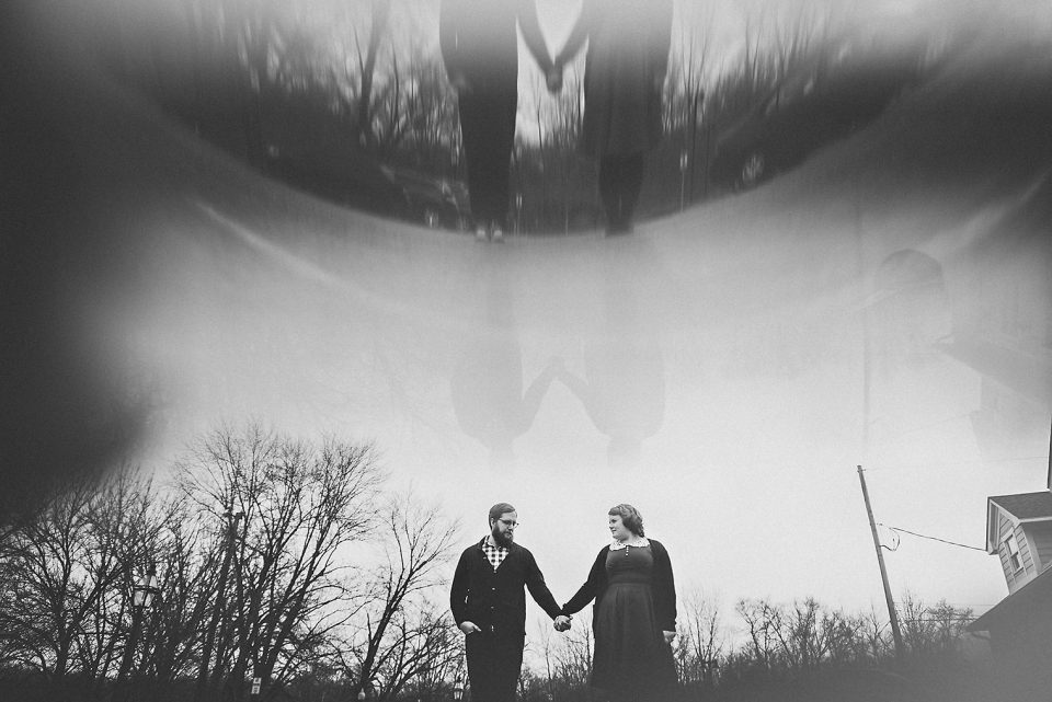 Monochrome photo of a couple holding hands, with a surreal, blurry reflection of their upper bodies in the sky through light refraction, surrounded by a misty landscape.
