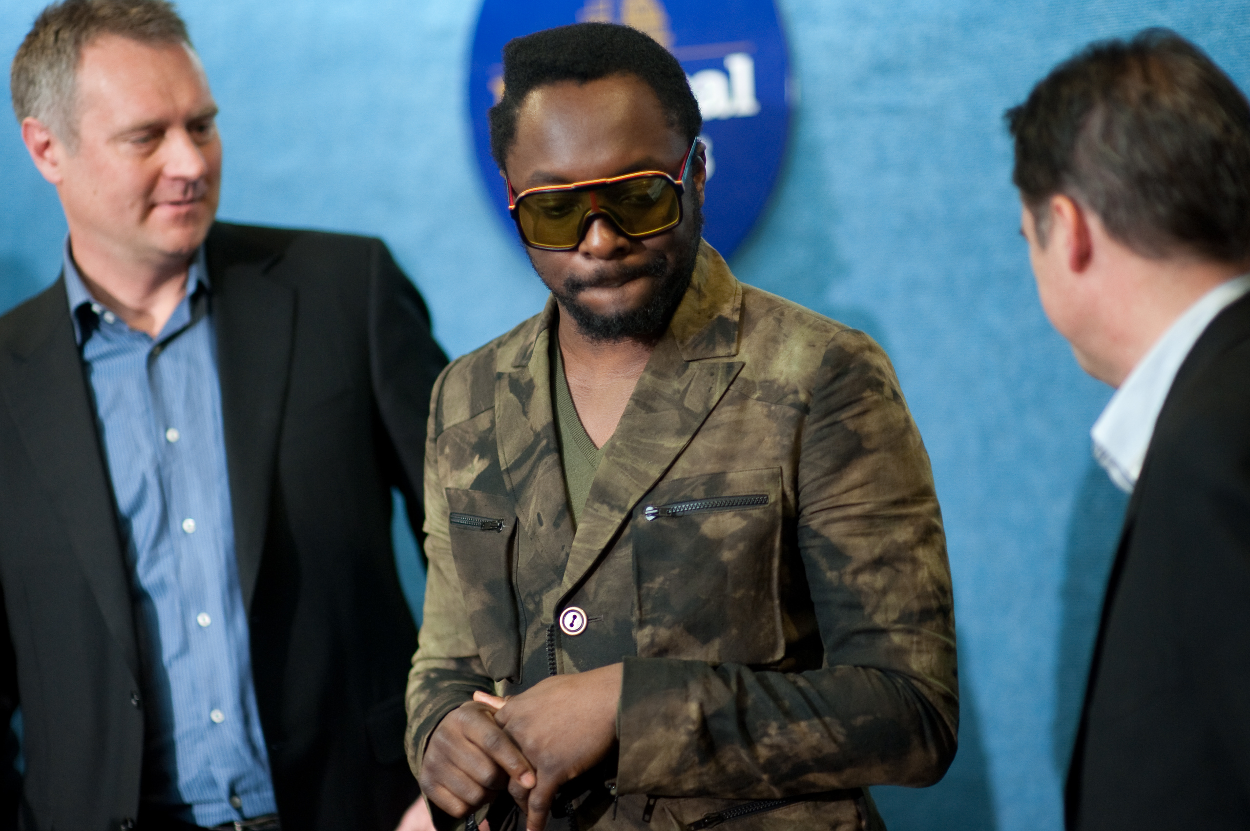 An epic portrait of a man in a camouflage jacket and sunglasses—resembling will.i.am—stands between two men in suits, set against a blue background with a circular emblem.