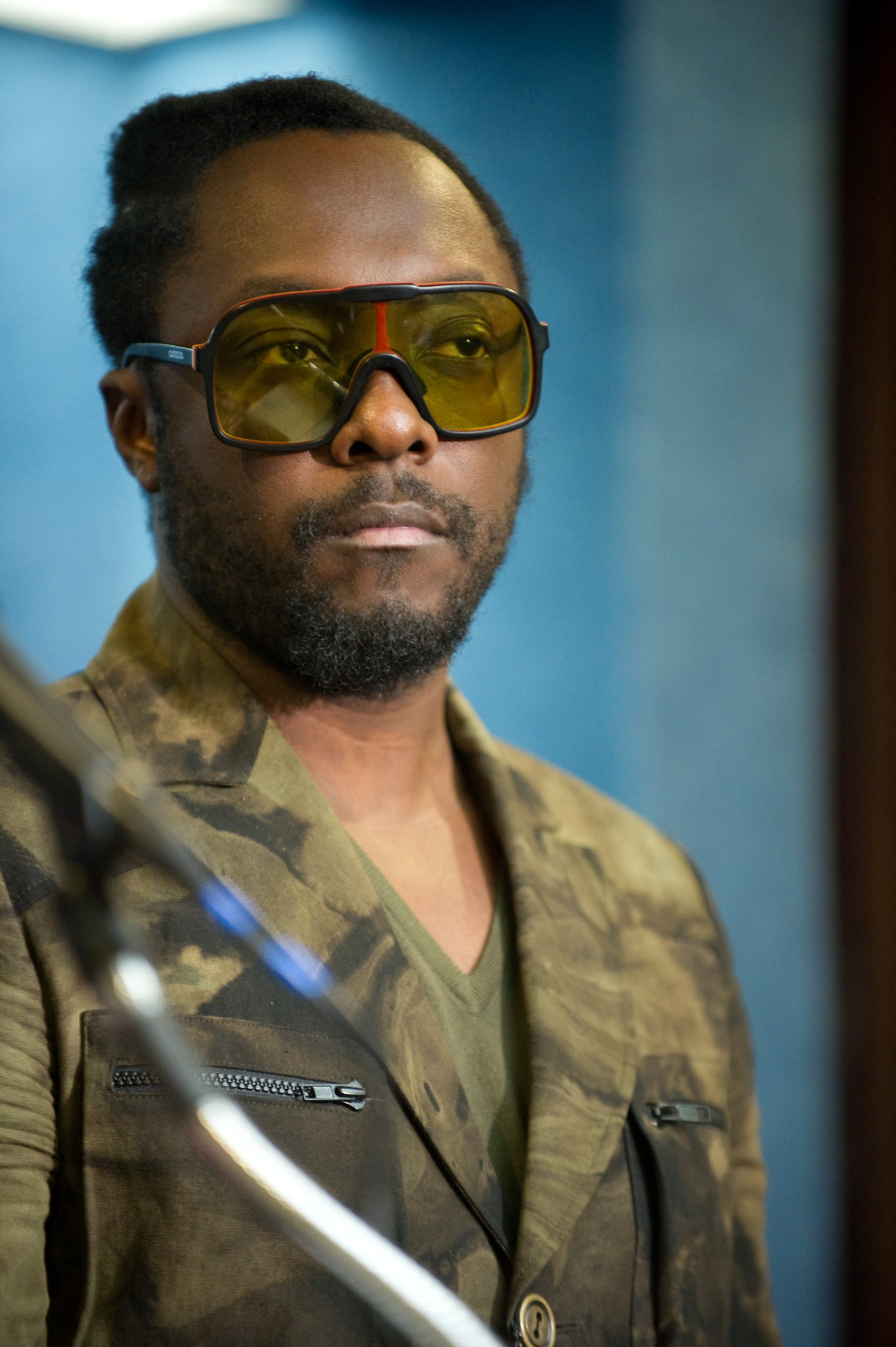 A man with a short beard and mustache wears large yellow-tinted glasses and a camouflage-patterned jacket, standing in front of a blue background—an epic portrait reminiscent of will.i.am’s bold style.