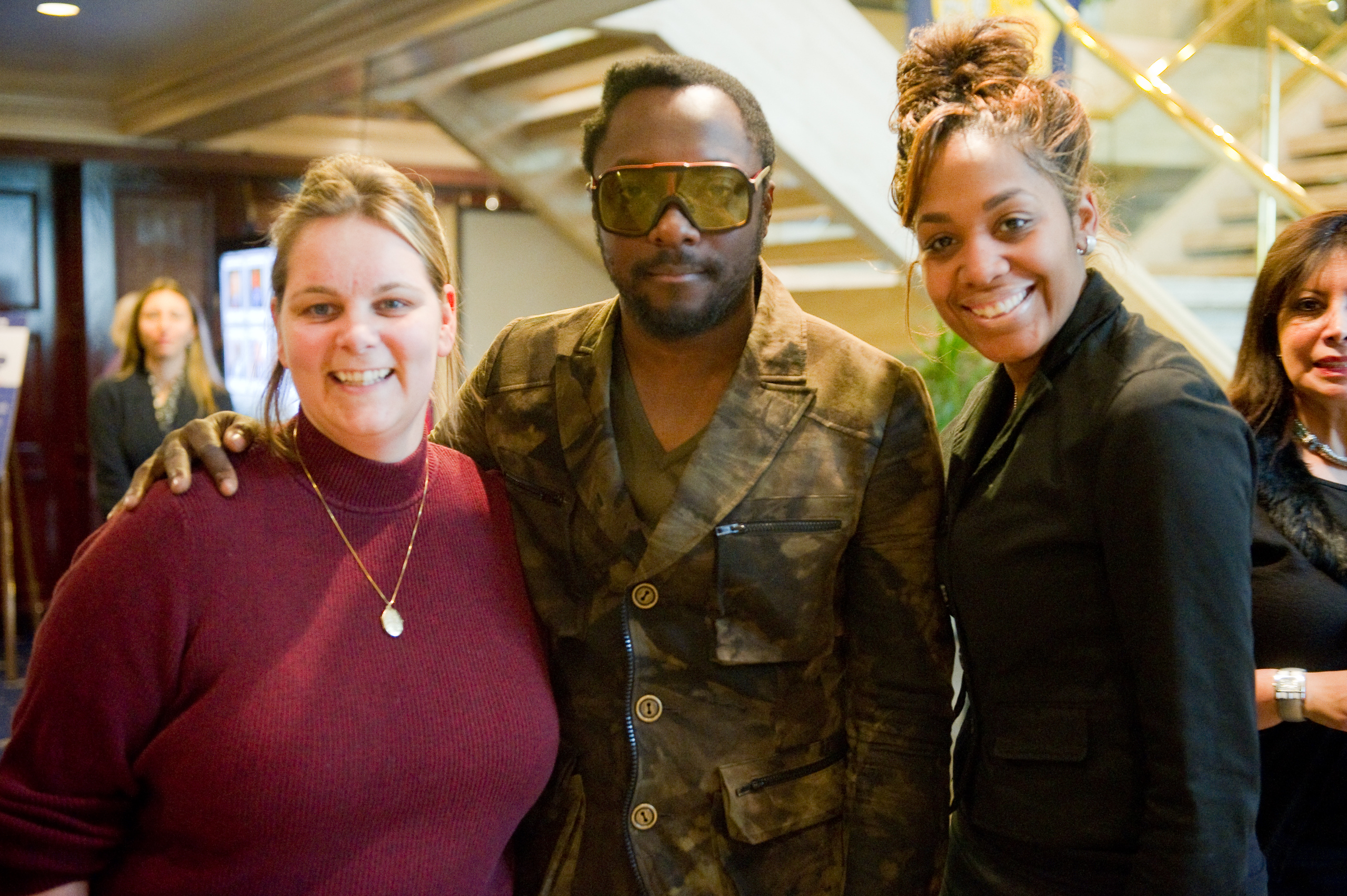 Three adults pose together indoors in this epic portrait—one wearing sunglasses and a camouflage-patterned jacket reminiscent of will.i.am, the others dressed in dark tops, all smiling at the camera.
