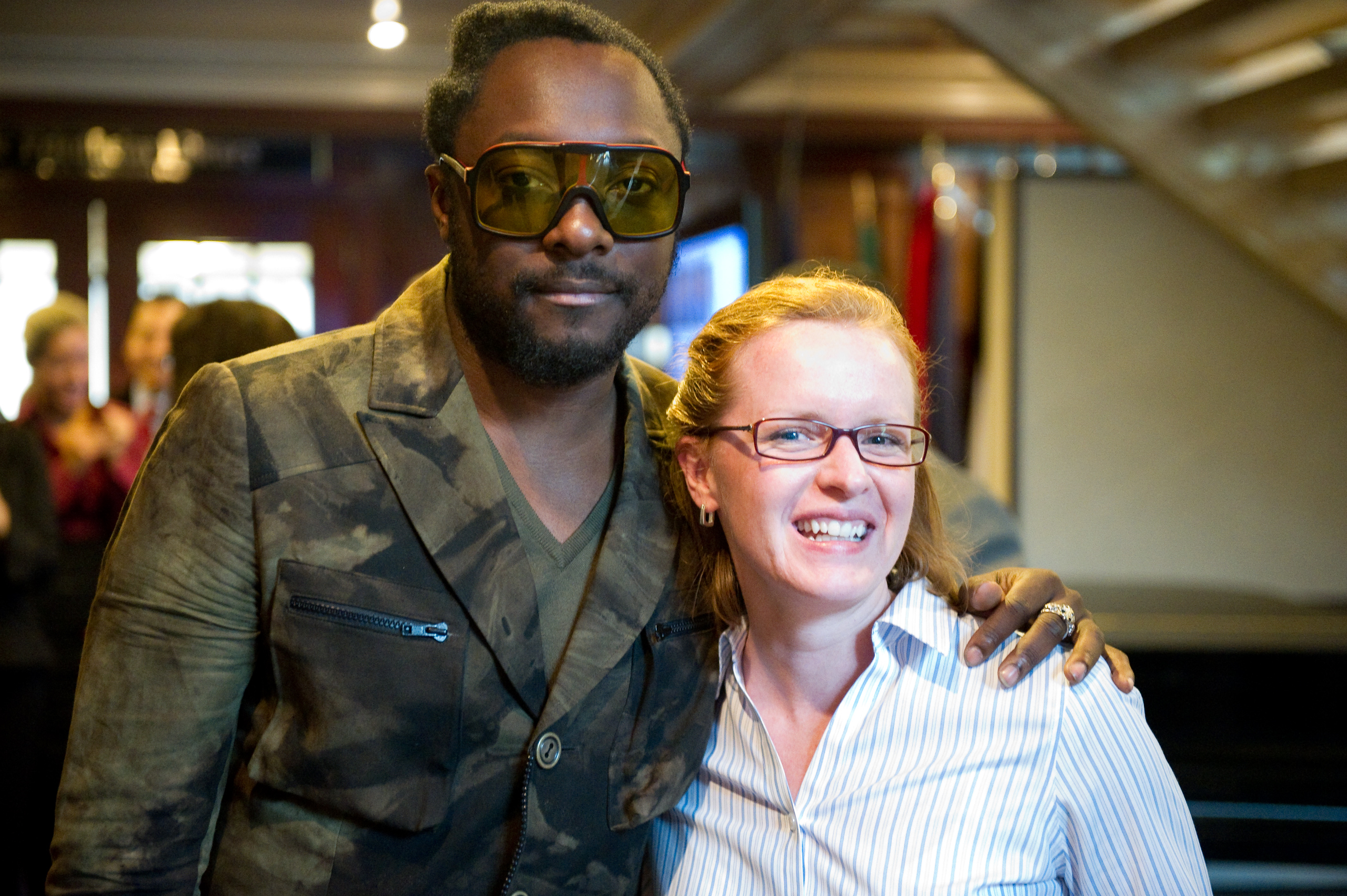 A man resembling will.i.am in sunglasses and a patterned jacket stands with his arm around a smiling woman in glasses and a striped shirt, creating an epic portrait as they pose indoors.