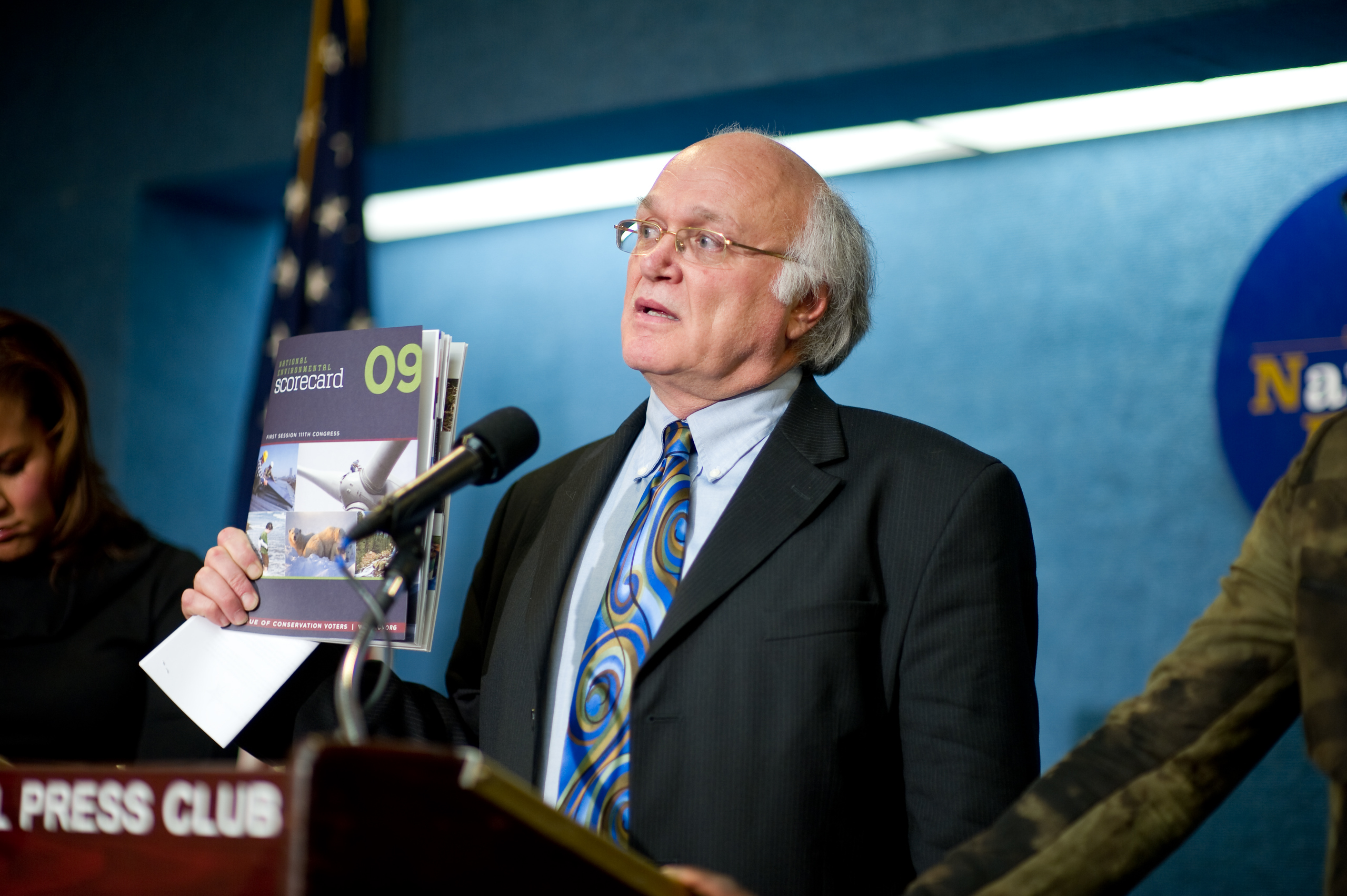An older man in a suit and patterned tie speaks at a podium, holding documents, with microphones and an epic portrait-style blue background behind him.