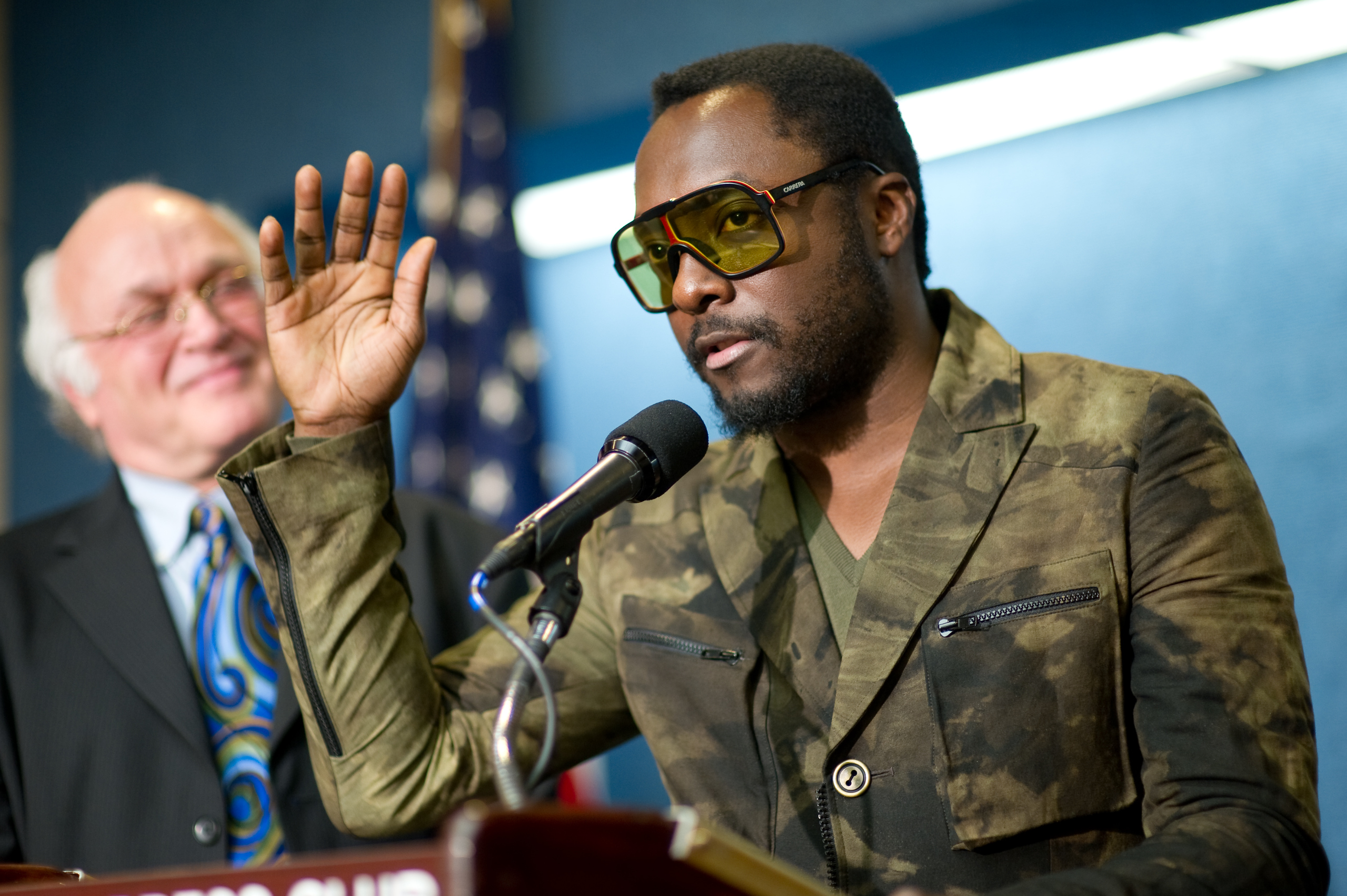 A man in camouflage-patterned clothing and yellow sunglasses delivers an epic portrait at the podium, while another man in a suit and tie, resembling will.i.am, stands behind him smiling.