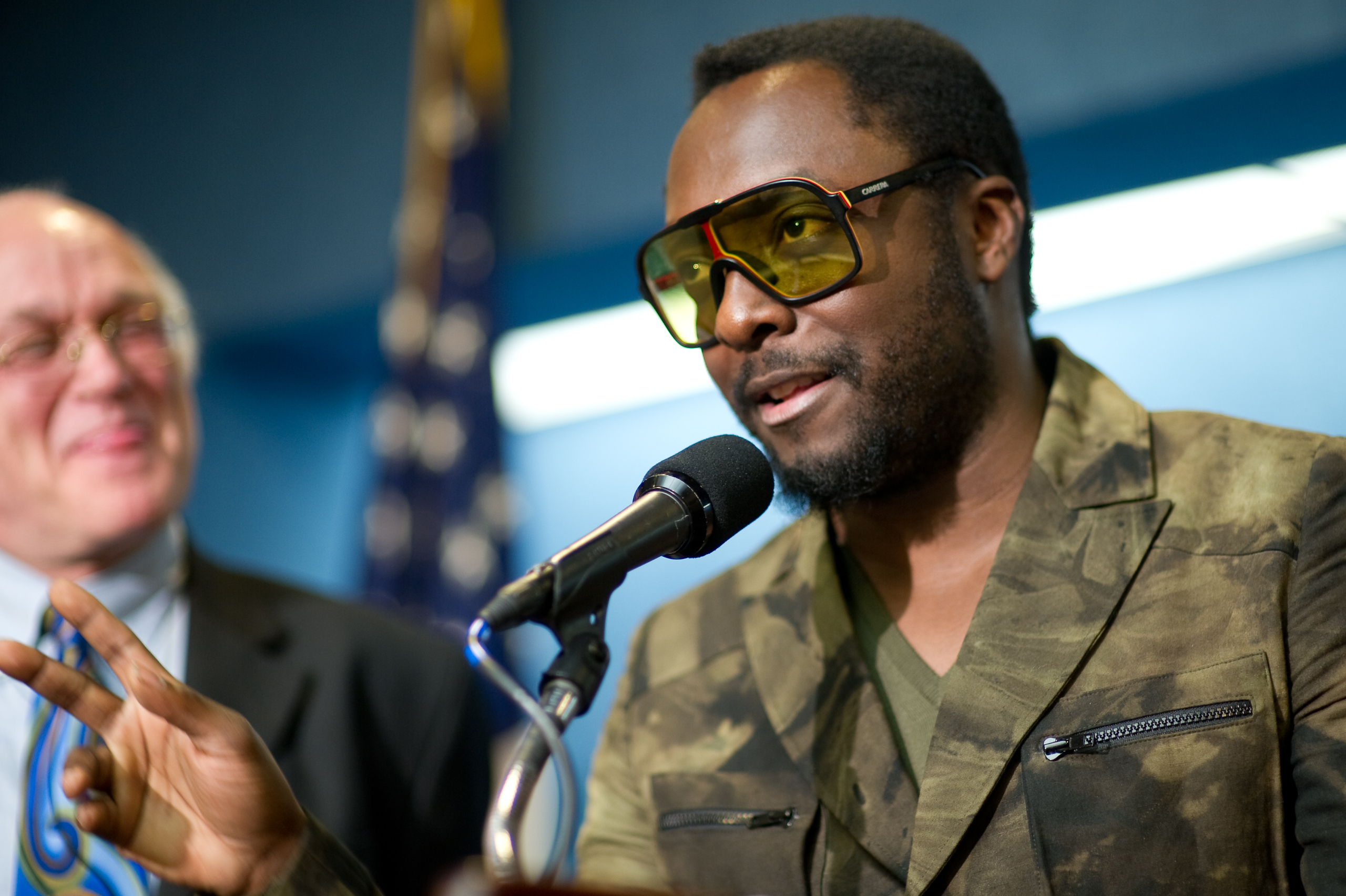 A man wearing large yellow glasses and a camouflage jacket—resembling an Epic Portrait of will.i.am—speaks into a microphone, gesturing with his hand; another man stands smiling in the background.