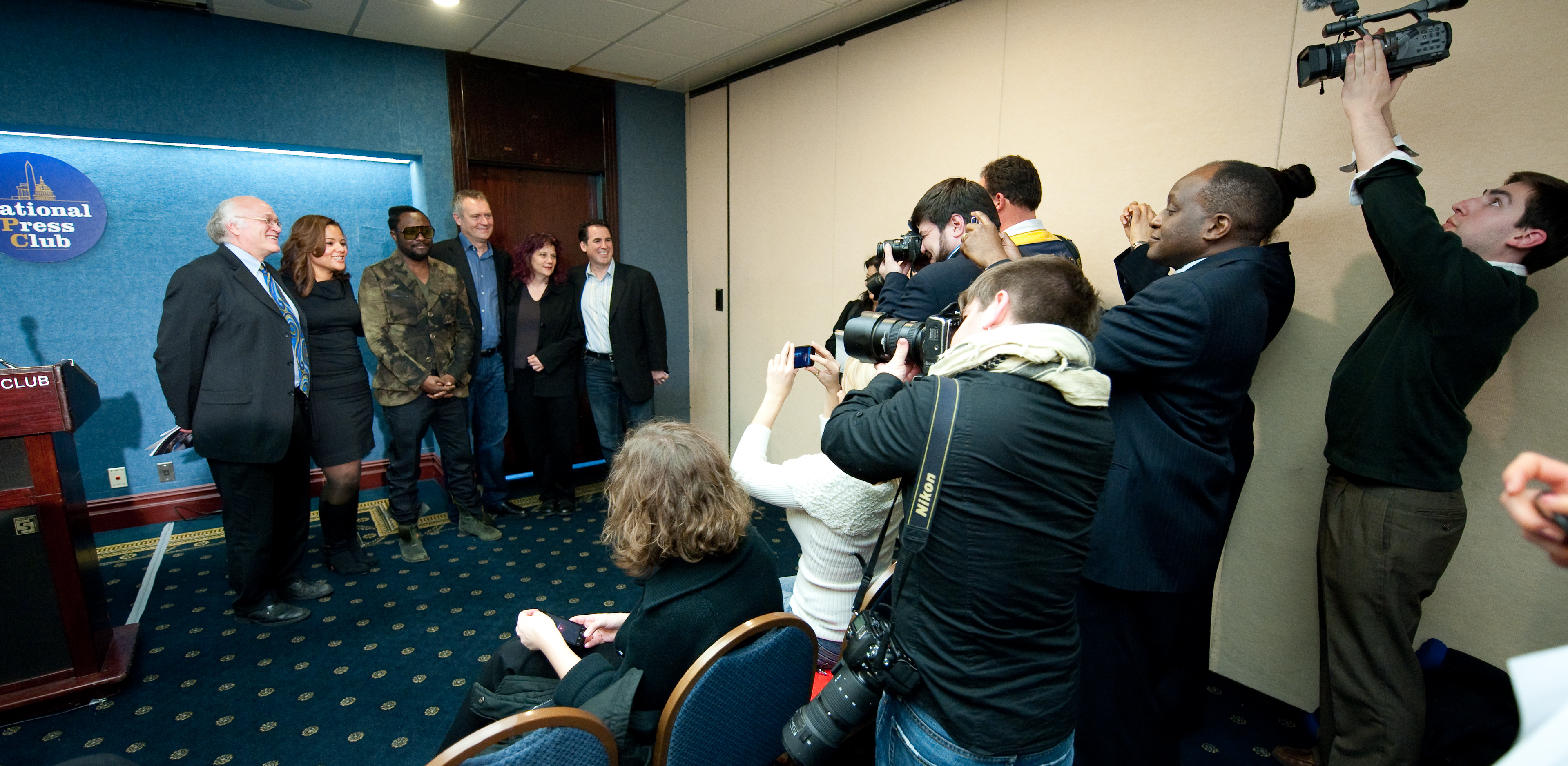 A group of people, including will.i.am, pose for an epic portrait at a press event as a crowd of journalists and photographers take pictures and record video.