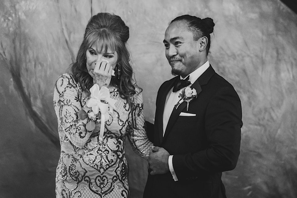 An emotional woman in a sequined dress wiping tears next to a smiling man in a tuxedo at an NYC wedding.