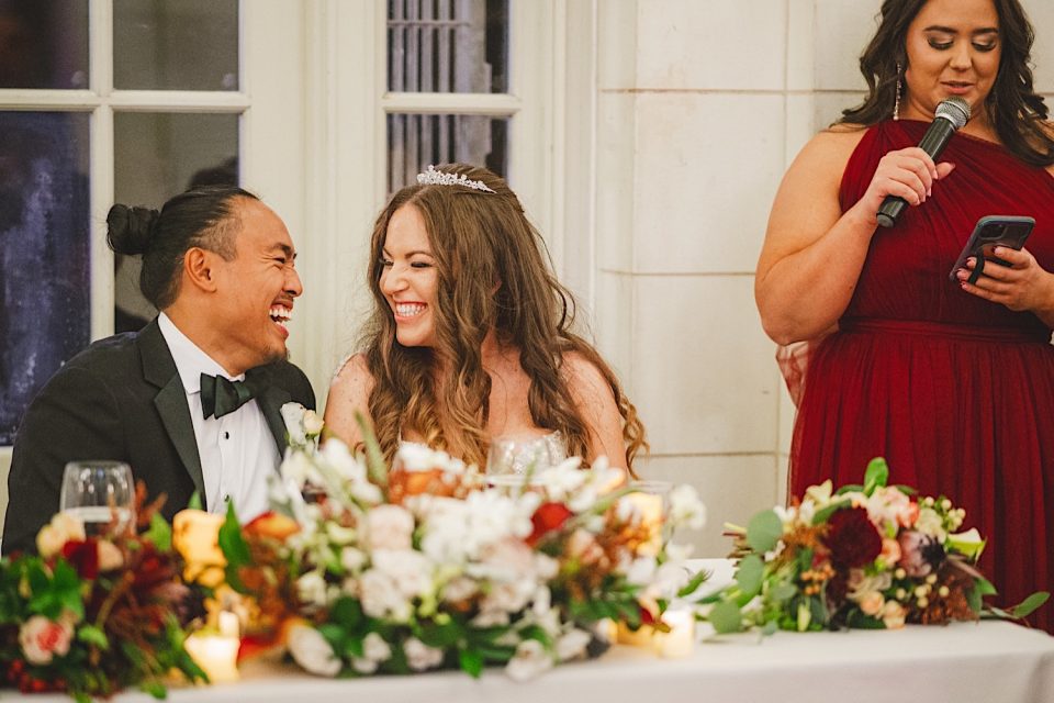 Ash and John laugh joyfully at their wedding table in the boathouse while a woman in a red dress gives a speech.