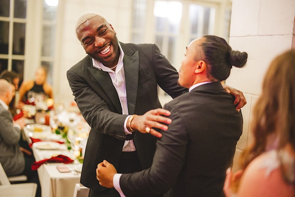 Two men joyfully embracing at an Ash + John Boathouse wedding reception, surrounded by guests seated at tables.