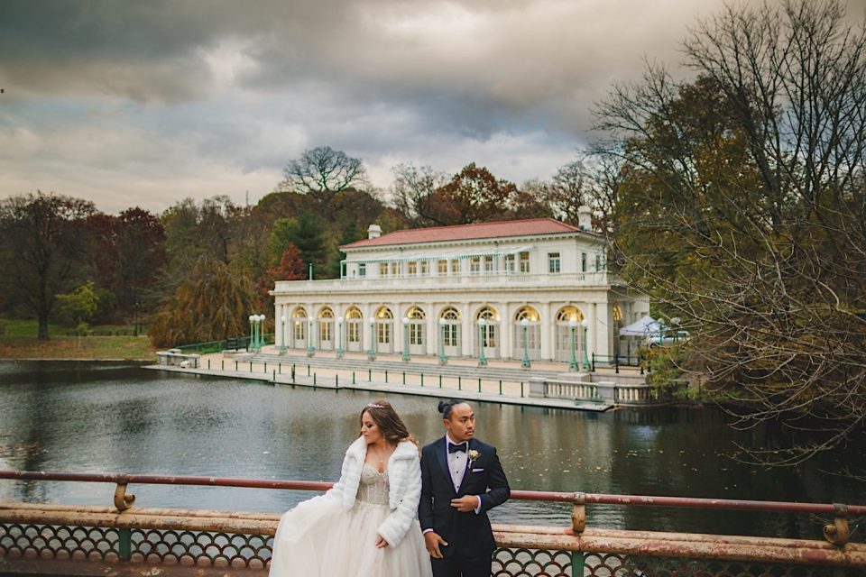 A bride in a white dress and a groom in a tuxedo walking by a lake with the Ash + John Boathouse in the background on a cloudy day.