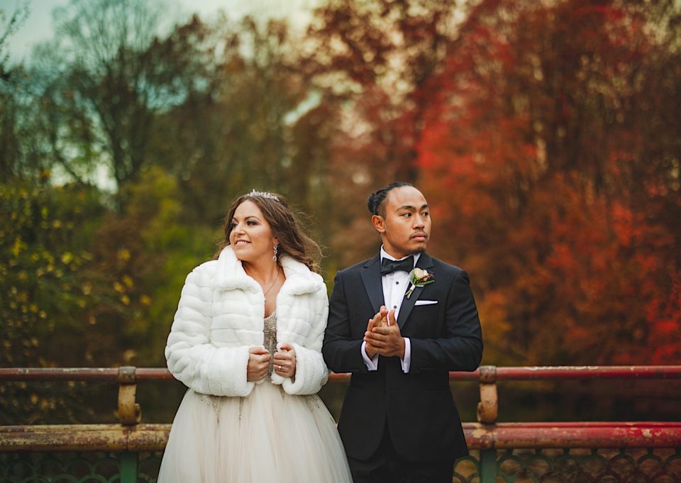 A bride in a white dress and fur coat stands with a groom in a black suit, holding hands on a bridge at the Ash + John Boathouse, with autumn trees in the background.