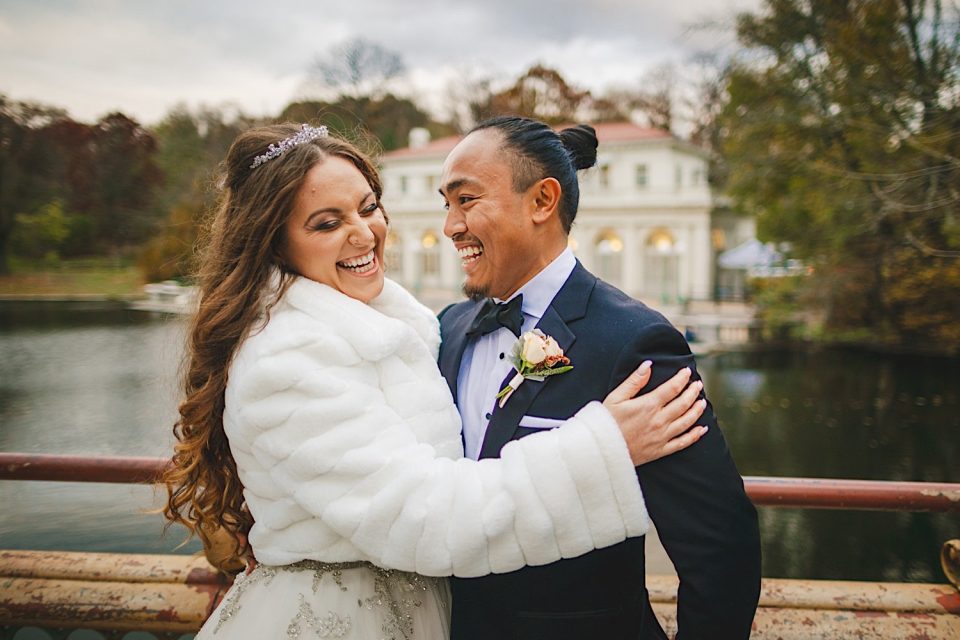 A joyful bride and groom embracing outdoors at their NYC wedding, the bride in a white dress and fur shawl, the groom in a black suit, with a scenic lake and gazebo in the background.