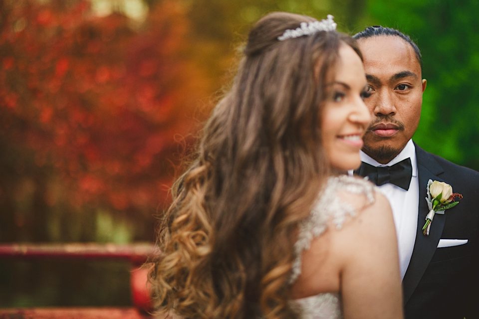Bride with curled hair and tiara smiles, standing partially out of frame at the NYC Wedding; behind her, a groom in a suit looks intently at the camera, with autumn trees in soft focus in the background.