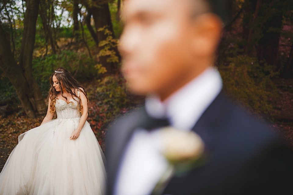 Bride in an elegant white dress looking down thoughtfully in a forest setting, with the groom out of focus in the foreground at Ash + John's NYC wedding.
