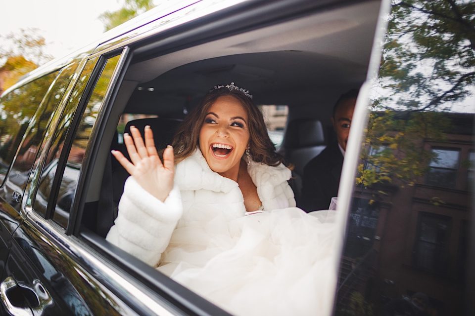 A joyful bride in a white dress and fur shawl waves from the backseat of a car, wearing a tiara, with a blurred NYC background.
