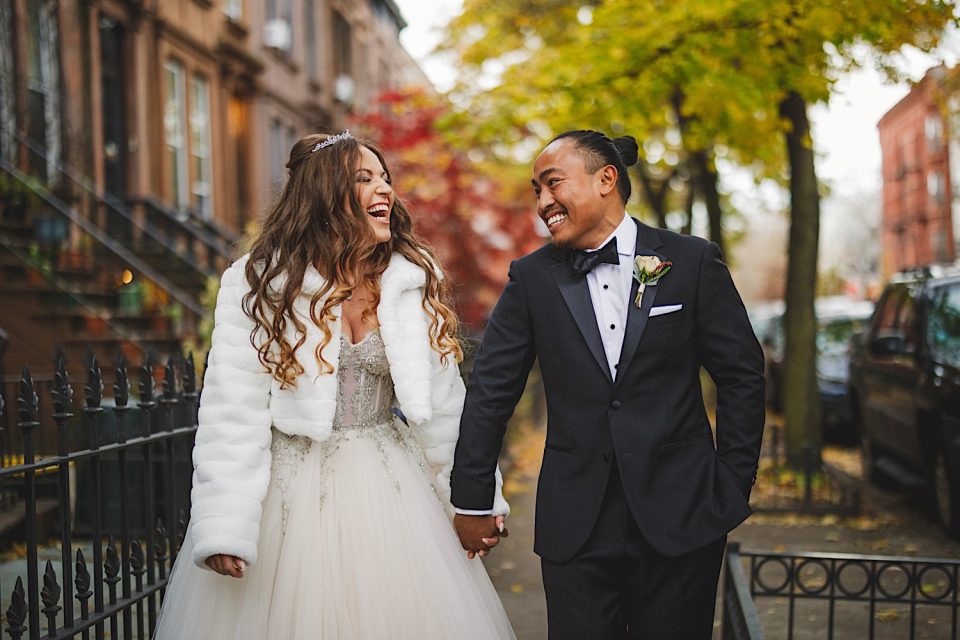 A joyful bride and groom holding hands and laughing on a city street lined with autumn trees during their NYC wedding.