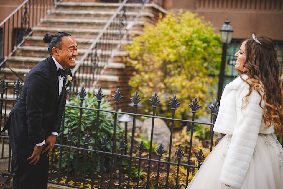 A bride and groom laugh joyfully outside on a leaf-strewn path by wrought iron fencing, with city brownstone steps in the background of their NYC wedding.