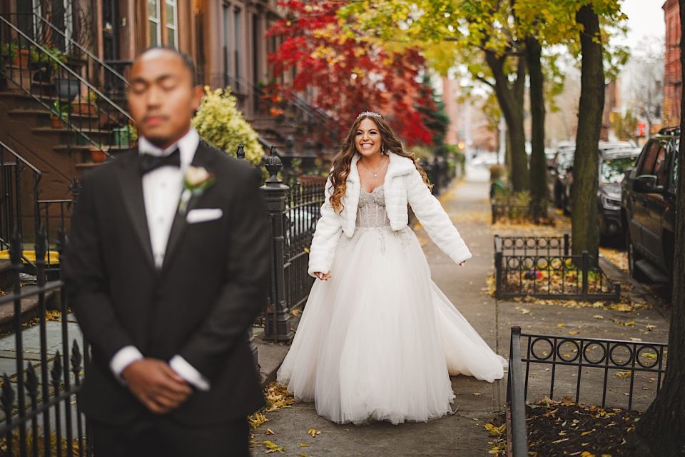 A bride in a white dress and fur jacket smiles joyfully as she approaches a groom in a black tuxedo, who is out of focus in the foreground, on a leaf-covered city street at their NYC Wedding.