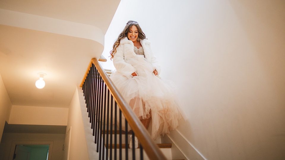 A joyful bride in a white gown and jacket descents a staircase at the John Boathouse, smiling broadly.
