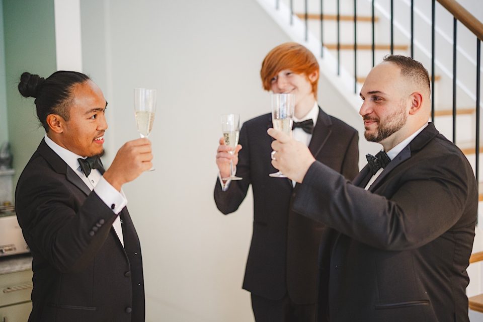 Three men in tuxedos, smiling and toasting with champagne glasses indoors at an Ash + John Boathouse NYC Wedding, with a staircase in the background.