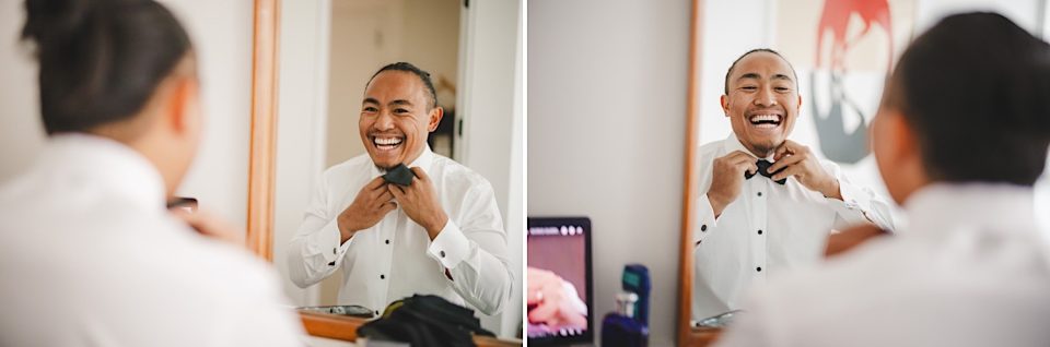 A joyful man tying his bowtie while laughing and looking at his reflection in a mirror at an NYC wedding.