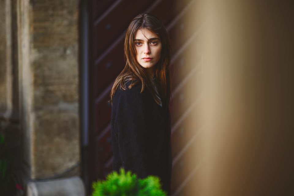 A woman with brown hair wearing a black coat, captured with a Nikon Z9, stands beside a building and looks towards the camera with a serious expression.