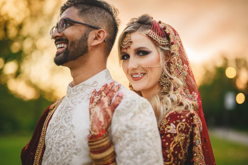 A joyful couple in traditional South Asian wedding attire, captured by the Nikon Z9, with the woman in a red lehenga and the man in a white sherwani, smiling outdoors at sunset.