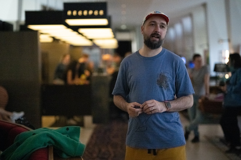 A man wearing a blue t-shirt and red cap stands indoors near a reception area, holding an object in his hands. People are visible in the background.