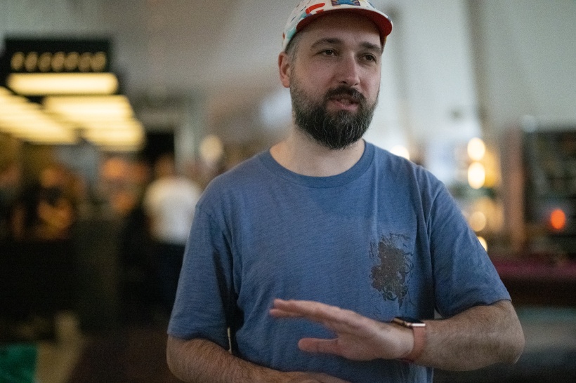 A bearded man wearing a blue T-shirt and a red and white hat gestures with his hand while speaking indoors. The background is slightly blurred.