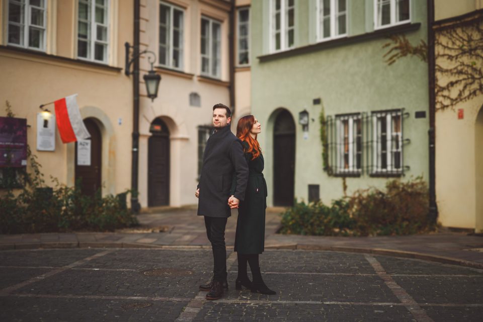 A man and woman stand holding hands on a cobblestone street in front of a row of colorful buildings, with a Polish flag visible in the background.