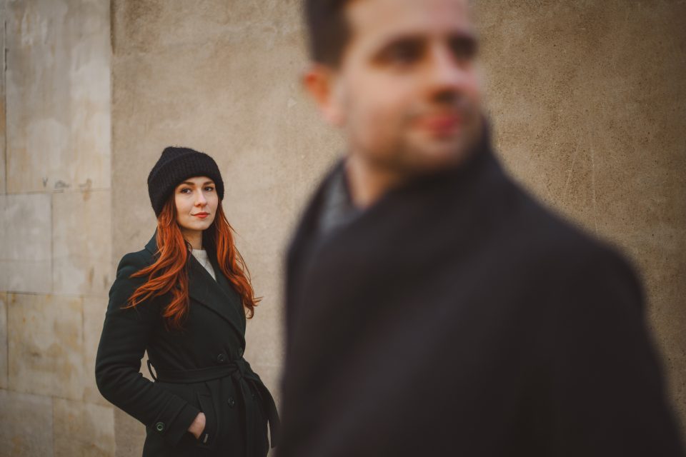 A woman in a black coat and hat stands against a stone wall, looking forward. A blurred man in a dark coat is in the foreground, partially facing her.