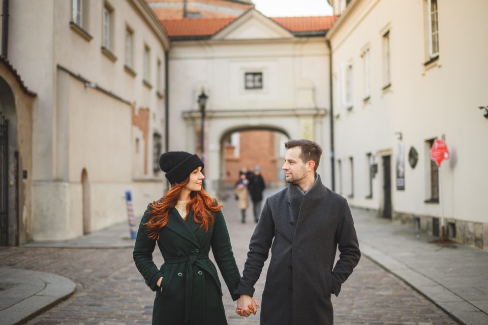 A couple holding hands and walking down a cobblestone street lined with historic buildings, both dressed in winter coats.