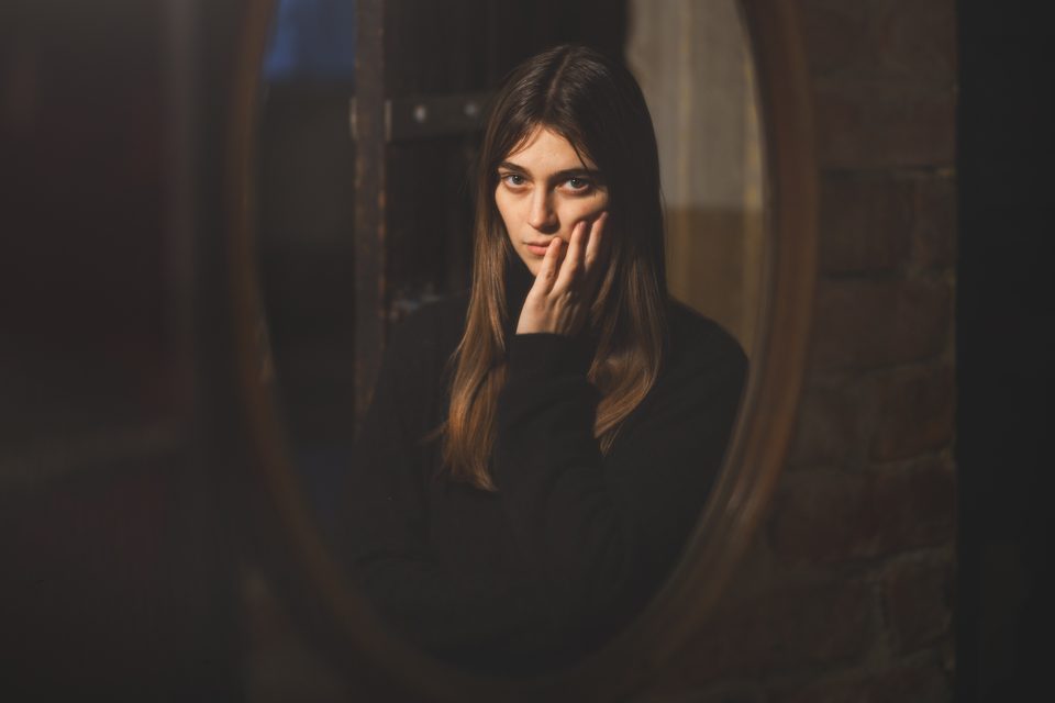 A woman with long brown hair, wearing a black top, looks into a round mirror with her hand touching her face. The background includes a partially opened door and a brick wall.